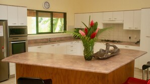 A kitchen with white cabinets, a wooden island, a clock on the wall, and a window with a view of greenery. - Mataheres Upstairs Muri (Muri Beach)