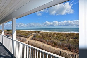 A beach view from a covered balcony with a wooden fence and a sandy path leading to the shore.