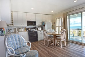 A compact kitchen with white cabinets, a stove, and a refrigerator. There is a dining table with four chairs and a view of the ocean through large windows.
