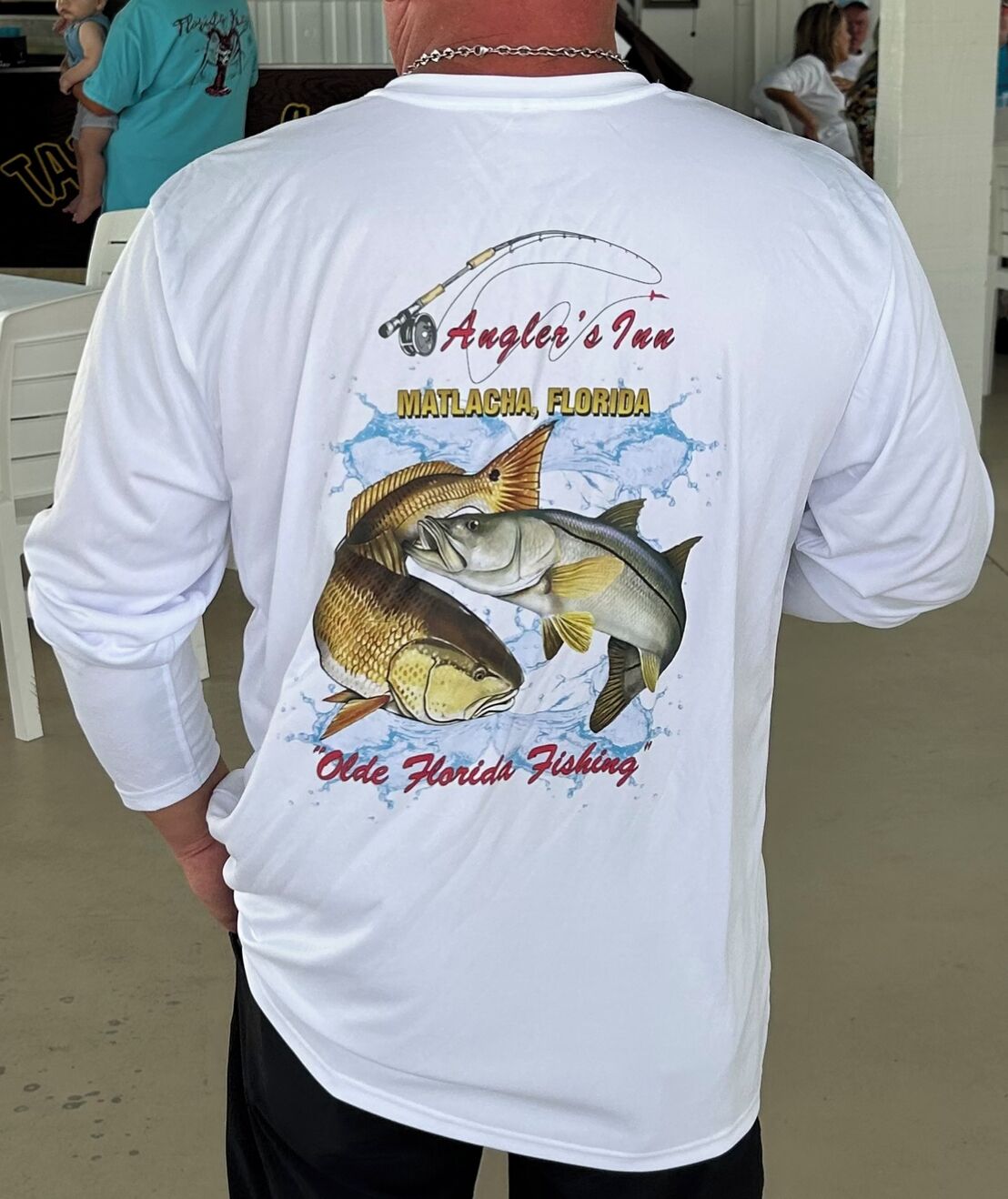 A white long-sleeve shirt with a fishing theme, featuring a fish and a fishing rod, and text indicating 'Angler's Inn Matlacha, Florida'.