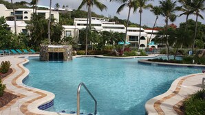 Une piscine agrémentée d’une cascade en pierre, entourée de palmiers et bordée d’un bâtiment blanc doté de balcons.