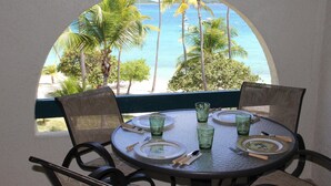 Une table ronde entourée de quatre chaises, dressée pour deux personnes, avec vue sur une plage bordée de palmiers et sur l’océan.