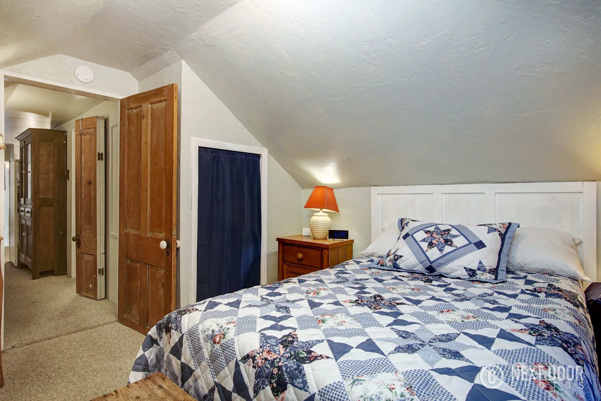A bedroom with a bed, a nightstand, a lamp, and a door leading to another room.
