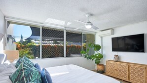 A bedroom with a bed, a ceiling fan, a TV, a window with a view of greenery, and a plant. - Burleigh Beach Pad 100m from beach - stunning (Burleigh Heads)