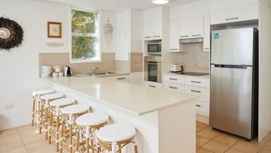 A modern kitchen with white cabinetry, a stainless steel refrigerator, a large island with stools, and a window with a view of greenery. - Burleigh Beach Pad 100m from beach - stunning (Burleigh Heads)