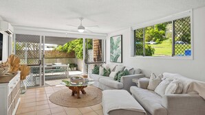 A spacious living area with a sofa, coffee table, and a view of a garden through large windows. - Burleigh Beach Pad 100m from beach - stunning (Burleigh Heads)
