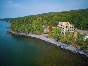 A large, multi-story house with a prominent roof, situated on a rocky shoreline with a forested hill in the background. - The Inn at Croftville Road Cottages #5. On the Shore of Lake Superior. (Grand Marais)