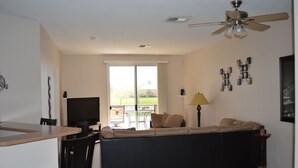 Ein Wohnzimmer mit einem dunklen Sofa, einem Fernseher, einem Deckenventilator und Blick durch ein großes Fenster.