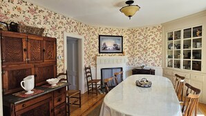 Une salle à manger comprenant une table, des chaises, une cheminée et un buffet dont le fond est recouvert d’un papier peint à motifs floraux.