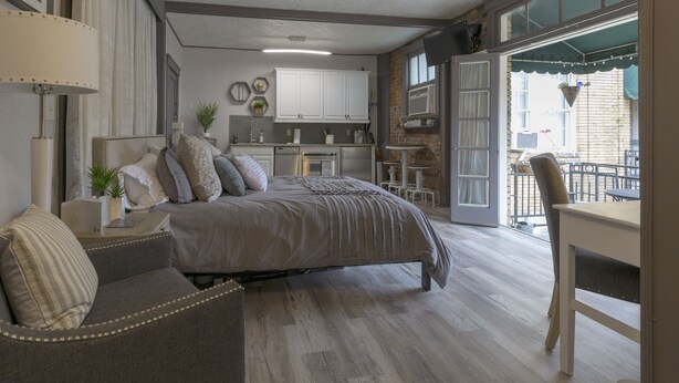A bedroom with a large bed, a sitting area with a chair, a dining table, and a kitchen area.