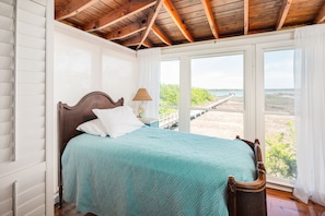 Een slaapkamer met een houten plafond, een bed met een blauwgroene spreidek, witte kussens en uitzicht op een strand en pier door grote ramen.