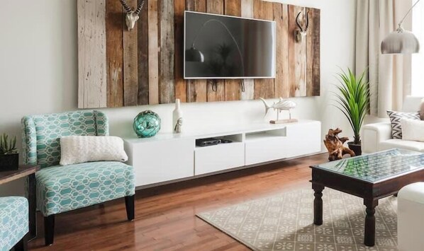 A living room with a wooden wall panel, a mounted flat-screen TV, a white sofa with patterned cushions, a coffee table, and a side table with decorative items.