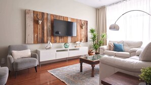 A modern living room with a flat-screen TV mounted on a wooden wall, a white sofa, a grey armchair, a coffee table, and a potted plant. - MTLVR #12 | Central, Kid-Friendly, Metro 3 min (Montreal)