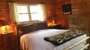 A wooden cabin bedroom with a bed, nightstands, a lamp, and a window with curtains. - Peaceful lakeside getaway (Meredith)