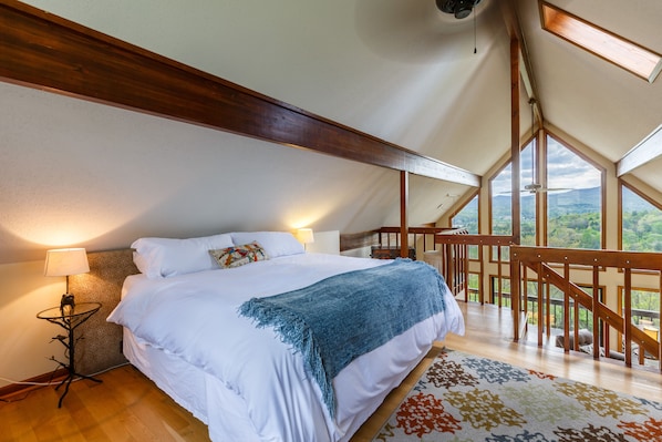 Ein Schlafzimmer mit einem großen Bett, einem Nachttisch, einer Lampe und Blick ins Freie durch ein großes Fenster.
