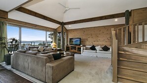 En rummelig stue med en stor sofa, en mindre sofa og et fjernsyn på en trævæg. Der er en trappe til højre og en balkon med udsigt.