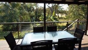 An outdoor dining area with a glass table and chairs, overlooking a lush garden with various plants and trees.