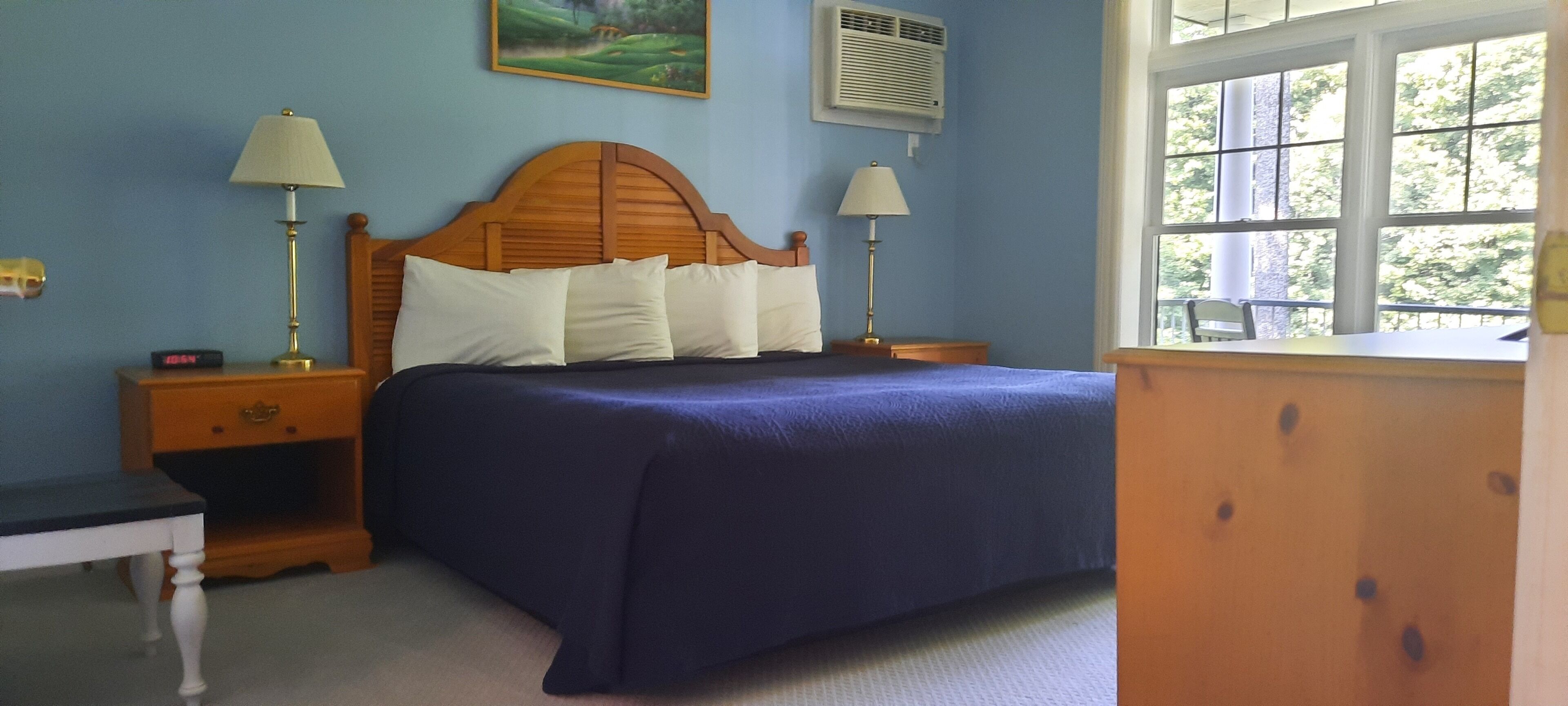 Une chambre à coucher avec une tête de lit en bois, une couvre-lit bleue, deux lampes de chevet, une table de chevet avec une horloge et une fenêtre donnant sur un paysage verdoyant.