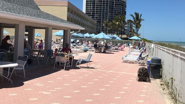 Eine Terrasse am Strand mit Sitzgelegenheiten im Freien, einem Poolbereich und einem hohen Gebäude im Hintergrund.