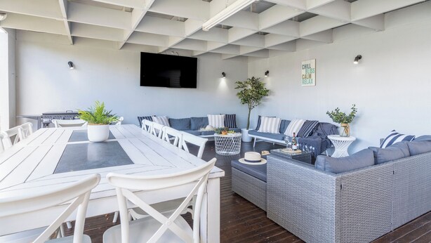Ein moderner Außenbereich mit Lounge-Möbeln, einem Esstisch, weißen Stühlen, einem grauen Sofa und einem an der Wand befestigten Fernseher.