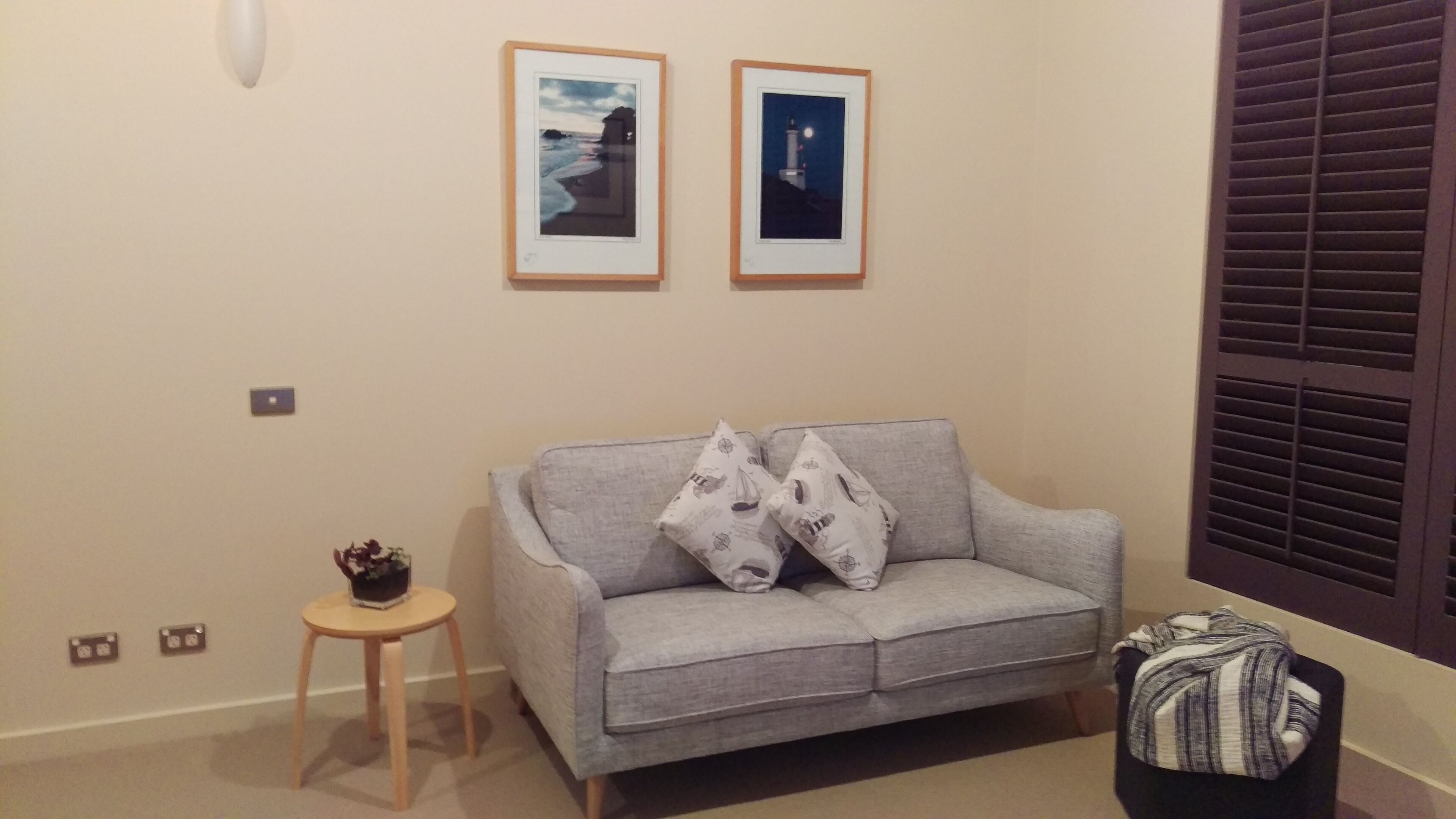 Een woonkamer met een grijze bank, twee ingelijste foto’s aan de muur, een klein houten krukje en een zwart voetenbankje.