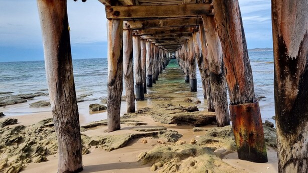 Un muelle que se adentra en el agua, con soportes de madera y una playa arenosa al final.
