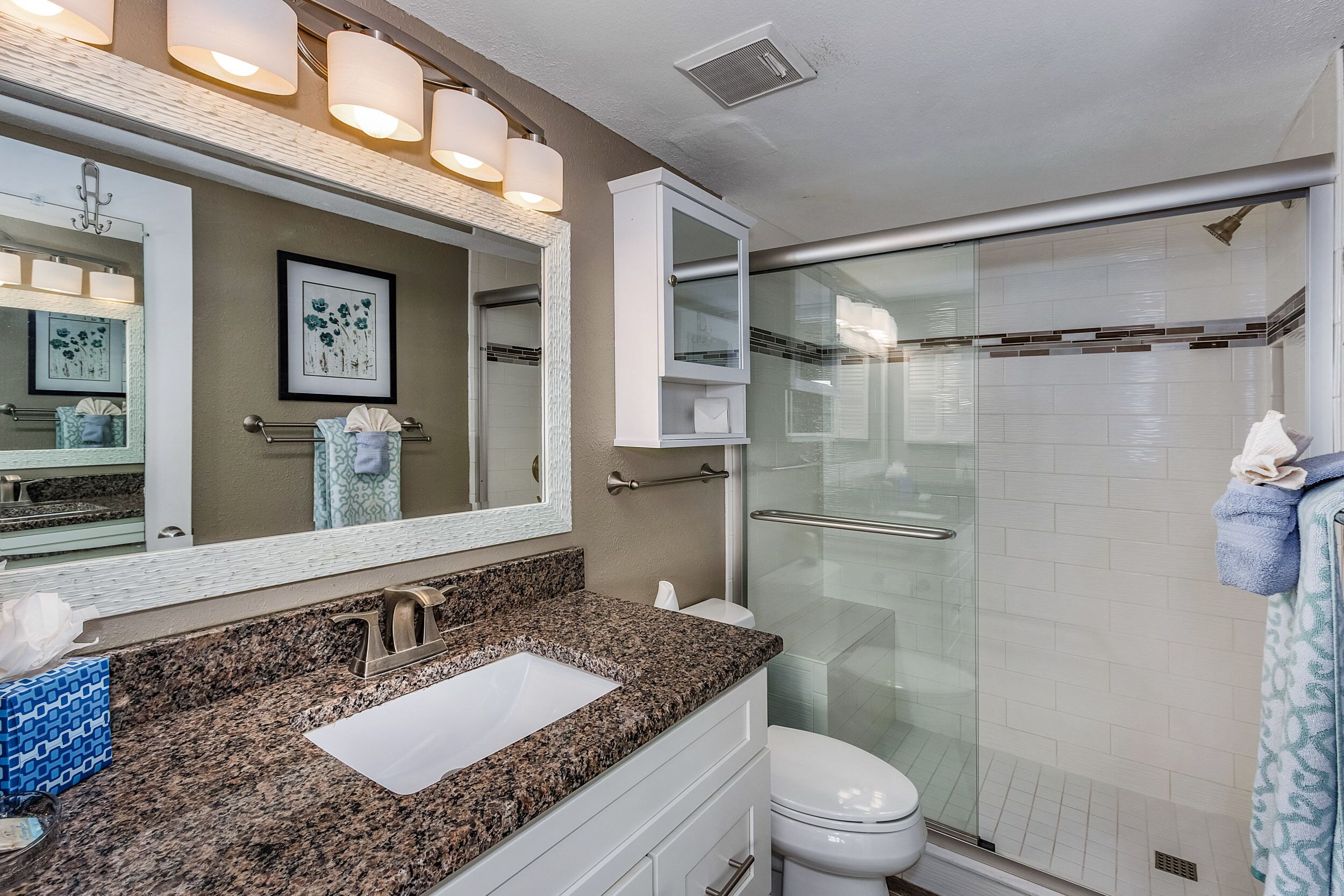 A bathroom with a granite countertop sink, a large mirror, a framed floral artwork, and a glass shower enclosure.