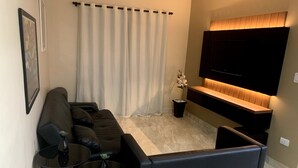 Un salon moderne avec un canapé d’angle, une table basse en verre, une petite table d’appoint, une plante en pot et un téléviseur à écran plat fixé au mur.