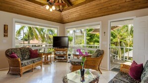 A spacious living room with a wooden ceiling, large windows, wicker furniture, and a flat-screen TV.
