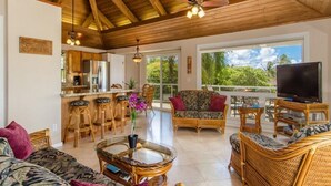 A spacious living area with a high ceiling, wooden beams, a kitchen with bar stools, a large TV, and a view of greenery outside.