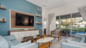 A modern living room with a large TV, light blue sofas, a coffee table, and a view of a swimming pool through large windows. - Luxury 5 Stars Home w/Princess & Mickey Themed Rooms, Game Room Private Pool/Spa (Kissimmee)