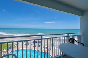 A balcony with a view of the ocean, a pool, and beach chairs.