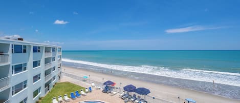A beachfront hotel with a pool, beach chairs, umbrellas, and a view of the ocean.