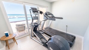 Ein Heimfitnessraum mit Laufband, Crosstrainer und Übungsball, ein kleiner Holztisch mit einer Wasserflasche sowie Blick auf einen Pool und einen Balkon.
