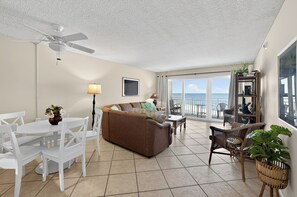 A living room with a brown sofa, white chairs, a dining table, a ceiling fan, and a view of the ocean through large windows.