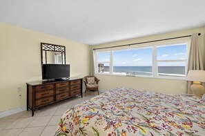 Un dormitorio con cama, cómoda, televisor, silla, ventana con vista al océano y lámpara.