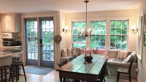 Un espace repas avec une table et des chaises en bois, un lustre et une vue sur la verdure grâce à de grandes fenêtres.