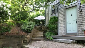 Un escalier en bois mène à une maison dont la porte est d’un vert clair, et qui est entourée d’un jardin agrémenté d’un muret en bois et de diverses plantes.
