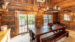 A rustic wooden cabin interior with a large dining table, benches, a chandelier, and a mounted deer head.