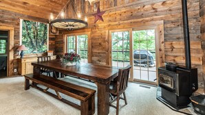 A rustic wooden interior with a large dining table, benches, a wood-burning stove, and large windows.