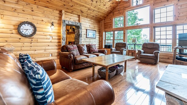 A cozy log cabin interior with a wooden ceiling, a stone fireplace, leather furniture, and large windows offering natural light.