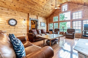 A cozy log cabin interior with a wooden ceiling, a stone fireplace, leather furniture, and large windows offering natural light.