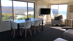 A room with a large dining table, white chairs, a TV, and a view of a lake and mountains. - SEA EAGLE COTTAGE Bay of Fires PEOPLES CHOICE TAS (Binalong Bay)