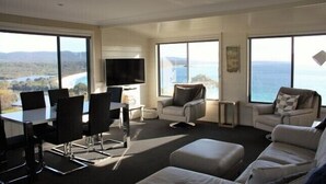 A spacious living room with a large dining table, comfortable seating, and large windows offering a scenic view of the ocean. - SEA EAGLE COTTAGE Bay of Fires PEOPLES CHOICE TAS (Binalong Bay)