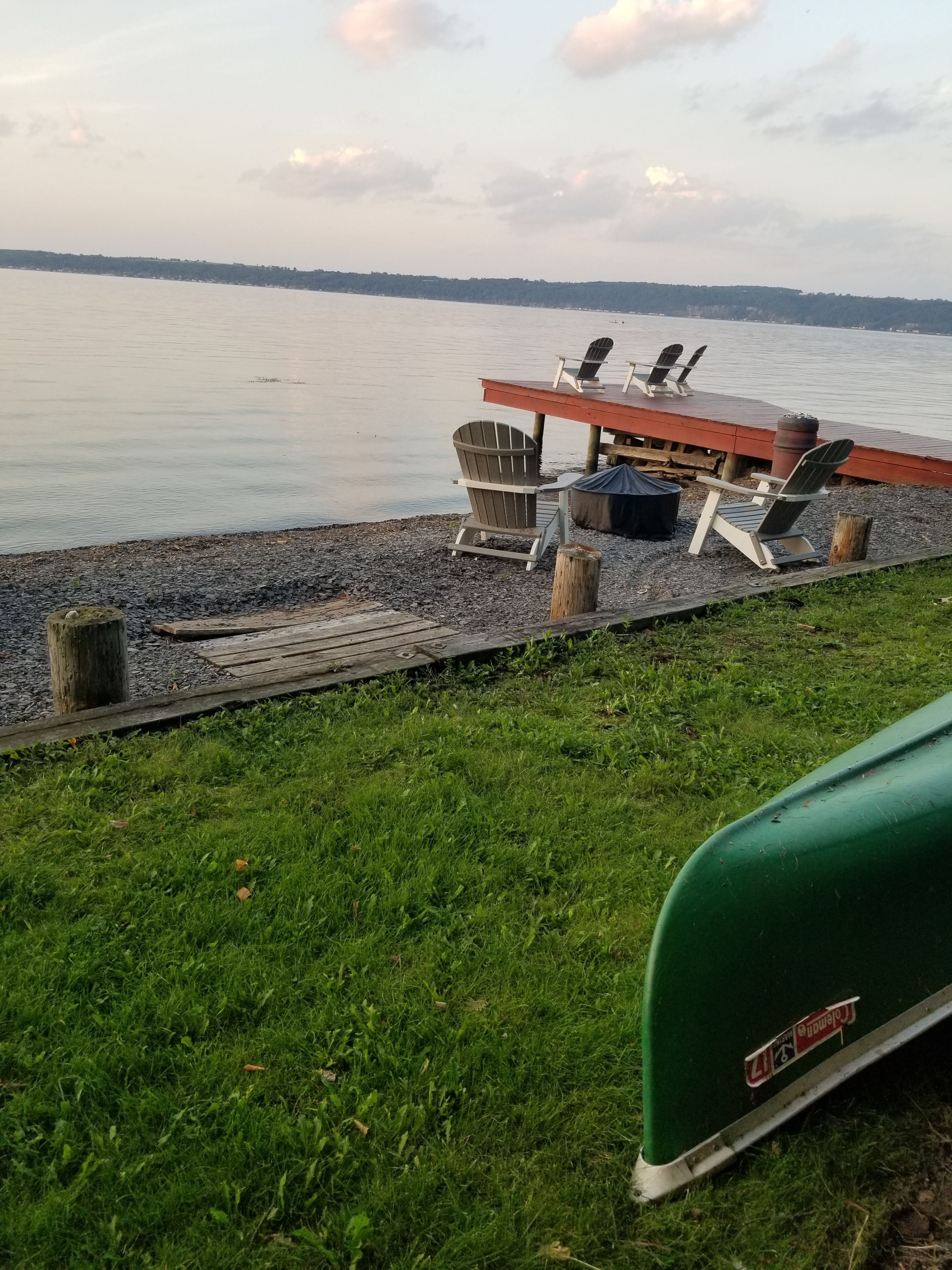 Terraza junto al lago con sillas Adirondack y una mesa, rodeada de césped y un muelle de madera.