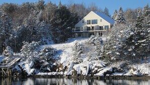 Ett stort hus i en våning med ett sluttande tak, omgivet av snötäckta träd och en sjö eller ett annat vattendrag.