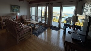 Ett rymligt vardagsrum med trägolv, en soffa, ett soffbord och ett skrivbord med en dator. Stora fönster ger utsikt över en småbåtshamn och havet.