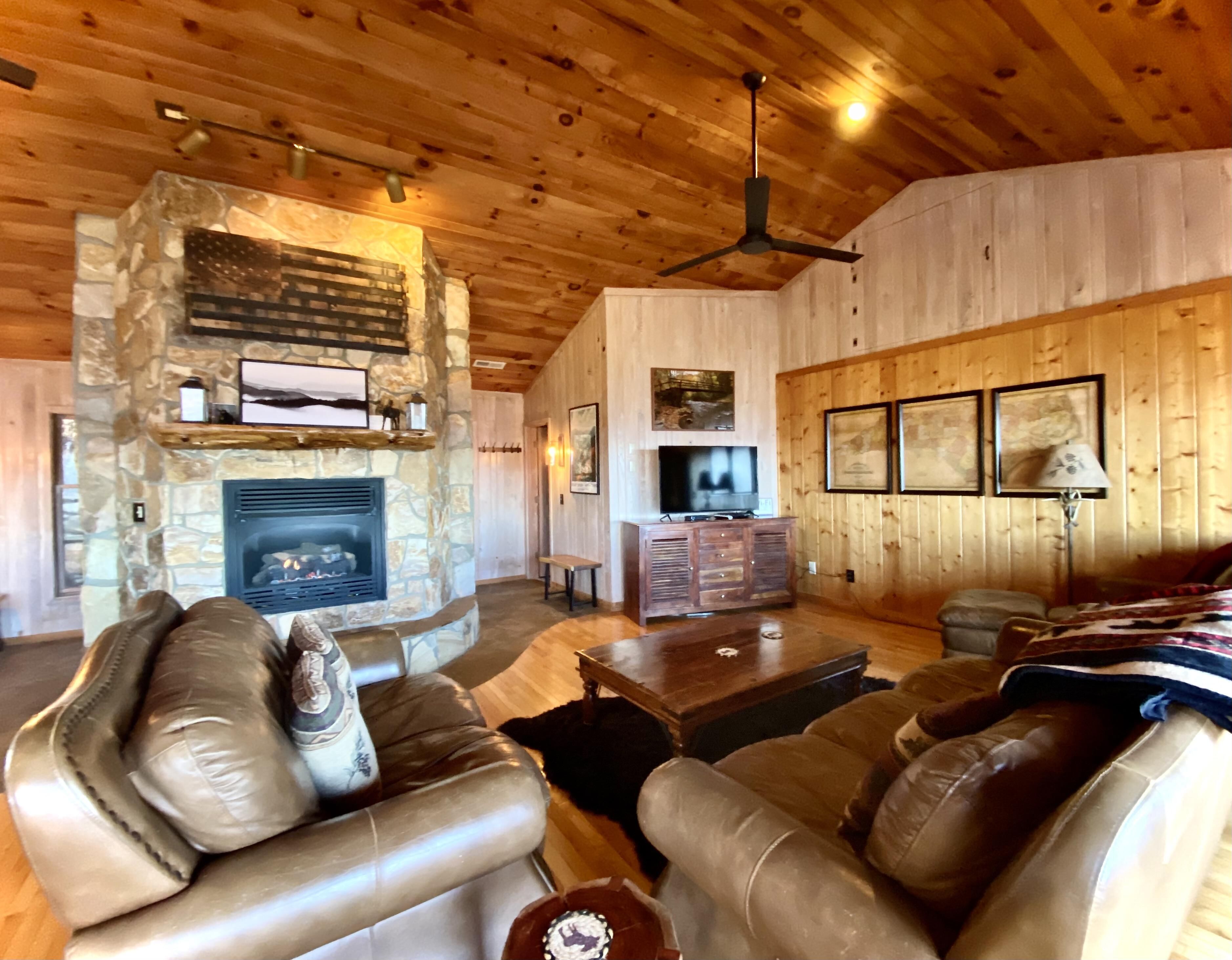 A cozy living room with a stone fireplace, leather sofas, a television, and wooden walls.