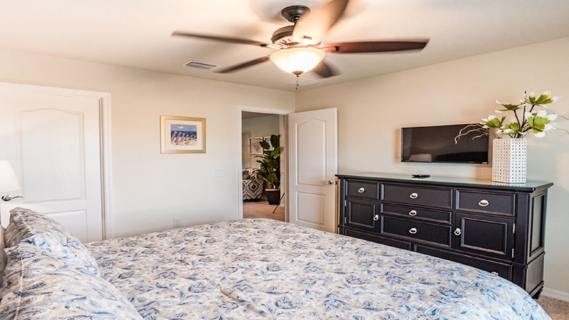 A bedroom with a bed, a dresser, a ceiling fan, a television, and a door leading to another room.