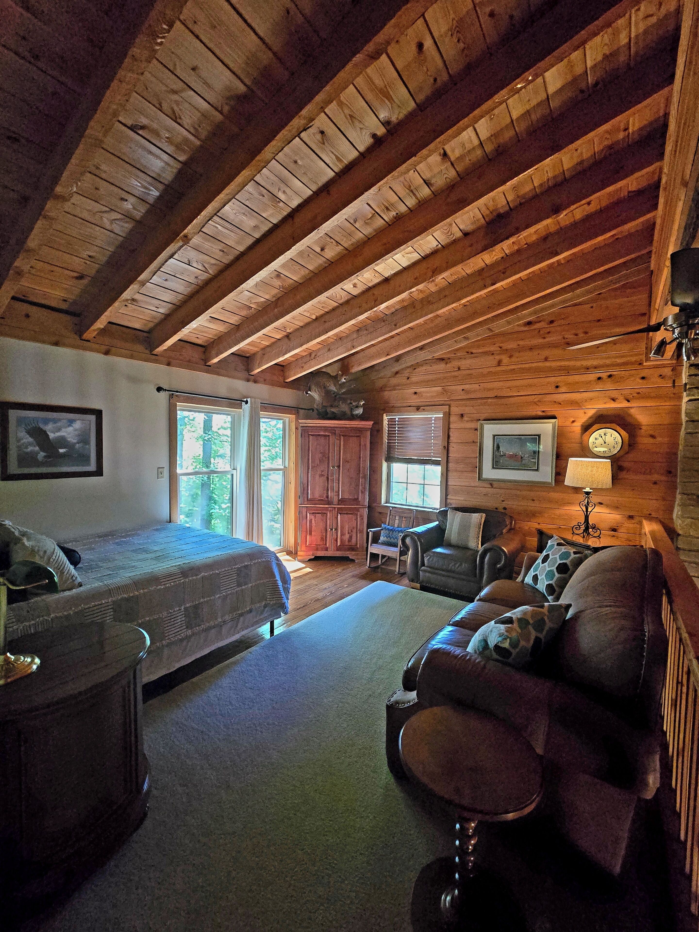 A log cabin interior with a bed, sofa, and armchair, wooden beams on the ceiling, and a clock on the wall.